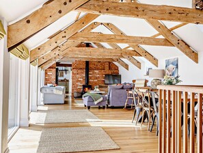 A spacious loft with exposed wooden beams, a brick fireplace, and a cozy seating area with cushions and a table.