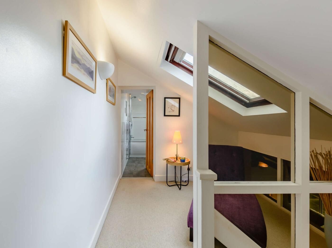 Un couloir avec un luminaire au plafond, une porte donnant sur une salle de bain et une petite table avec une lampe.
