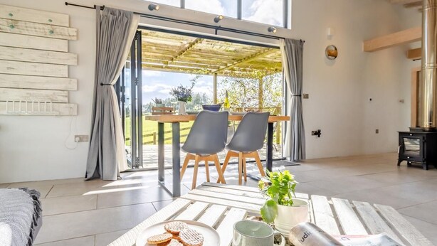 A modern room with a large window, a dining table with chairs, a fireplace, and a cozy seating area with a rug and cushions. - 1 Bed in Llandrindod Wells (oc-53194) (Llandrindod Wells)