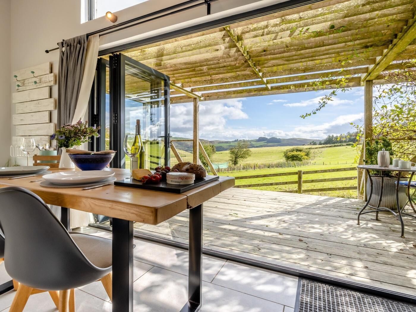 A dining area with a table set for two, a view of a green landscape, and a pergola structure.