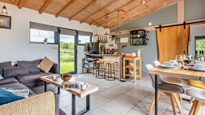 Una cocina moderna con vigas de madera, un comedor con mesa y sillas, un sofá y un ventanal con vistas a zonas verdes.