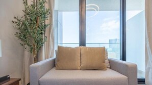 En moderne stue med en sofa, en potteplante og udsigt over byen gennem store vinduer.