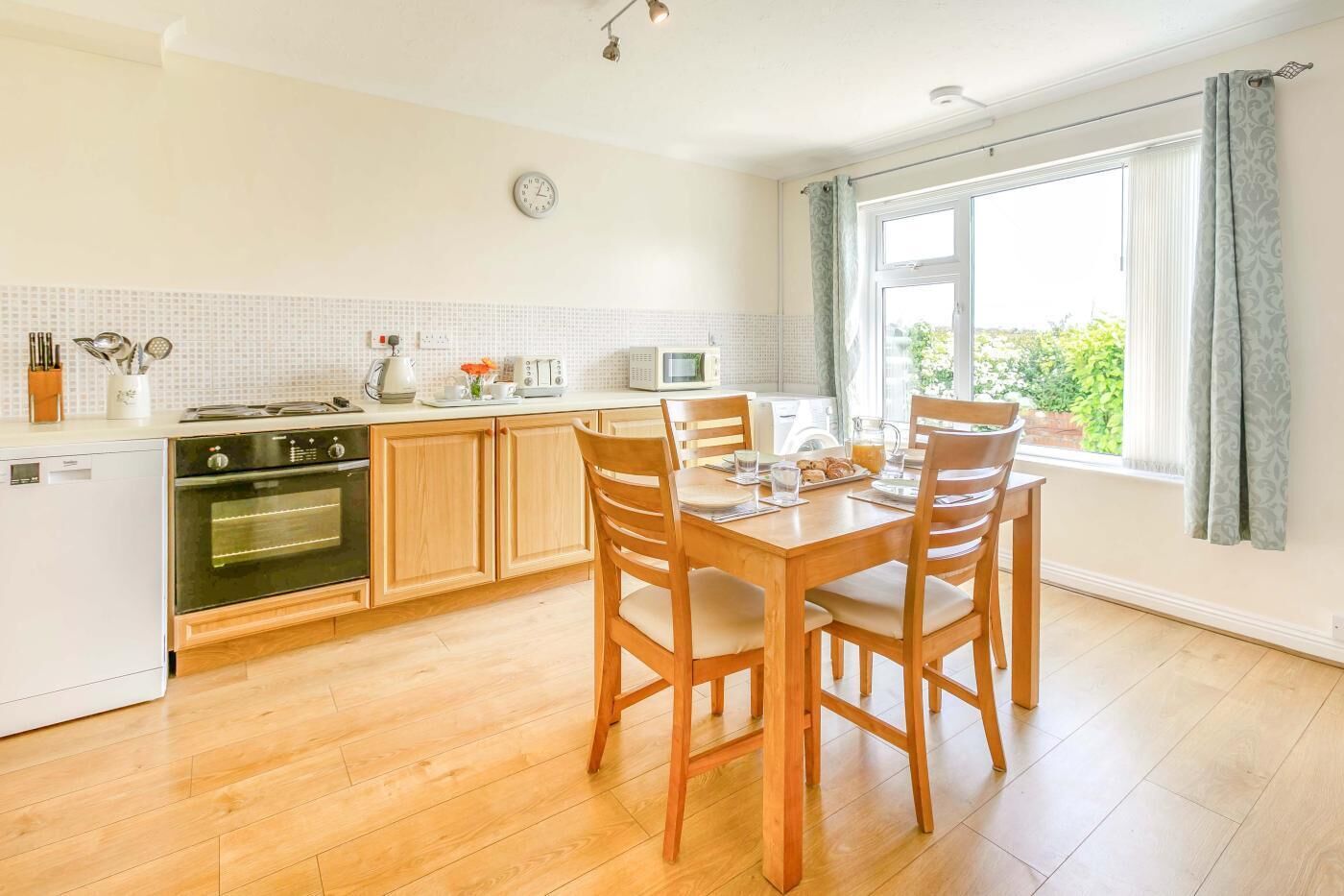 A kitchen with wooden cabinets, a dining table with chairs, a microwave, and a clock on the wall.