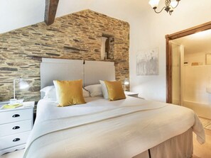 Un dormitorio con pared de piedra, una cama con ropa de cama blanca y dos almohadas amarillas, una mesita de noche con lámpara, y una puerta de madera.