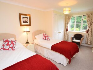 A bedroom with two beds, a nightstand with a lamp, a chair, a window with curtains, and a picture on the wall. - 2 Bed in Sherborne (oc-53673) (Chetnole)