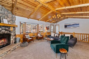 A cozy living room with a stone fireplace, wooden ceiling beams, and a view of the outdoors through large windows. - Big Bear Ski Chalet - Walk to Slopes & Hot Tub (Big Bear Lake)