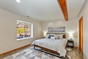 Quarto com cama, duas mesas de cabeceira, janela e teto com vigas aparentes.