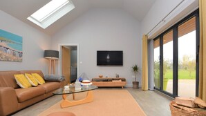 Ein geräumiges Wohnzimmer mit hohen Decken, einem an der Wand befestigten Flachbildfernseher, einer Sofagarnitur, einem Couchtisch und einem großen Fenster mit Blick ins Freie.
