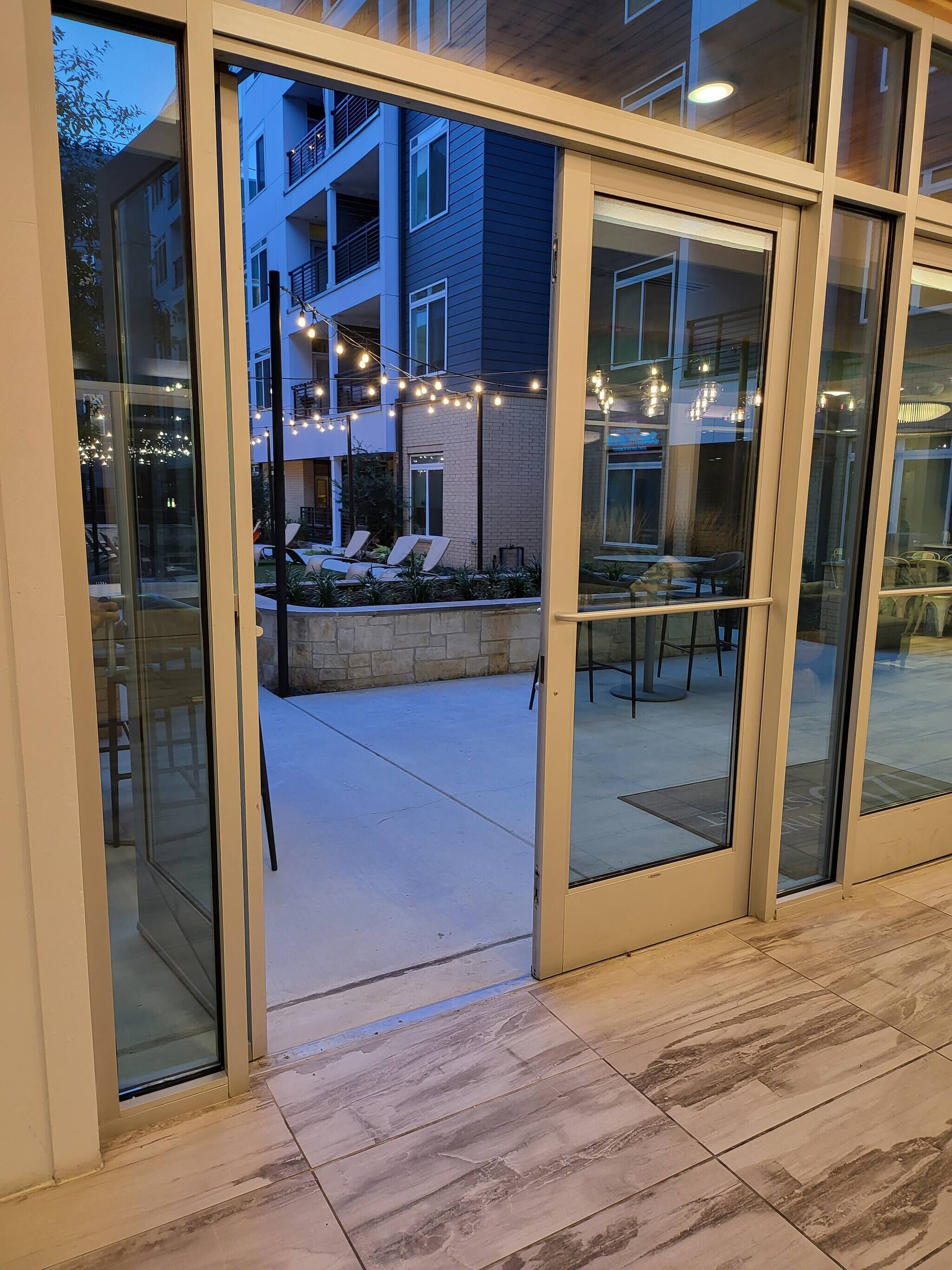 A glass door entrance to a building with outdoor seating and string lights.