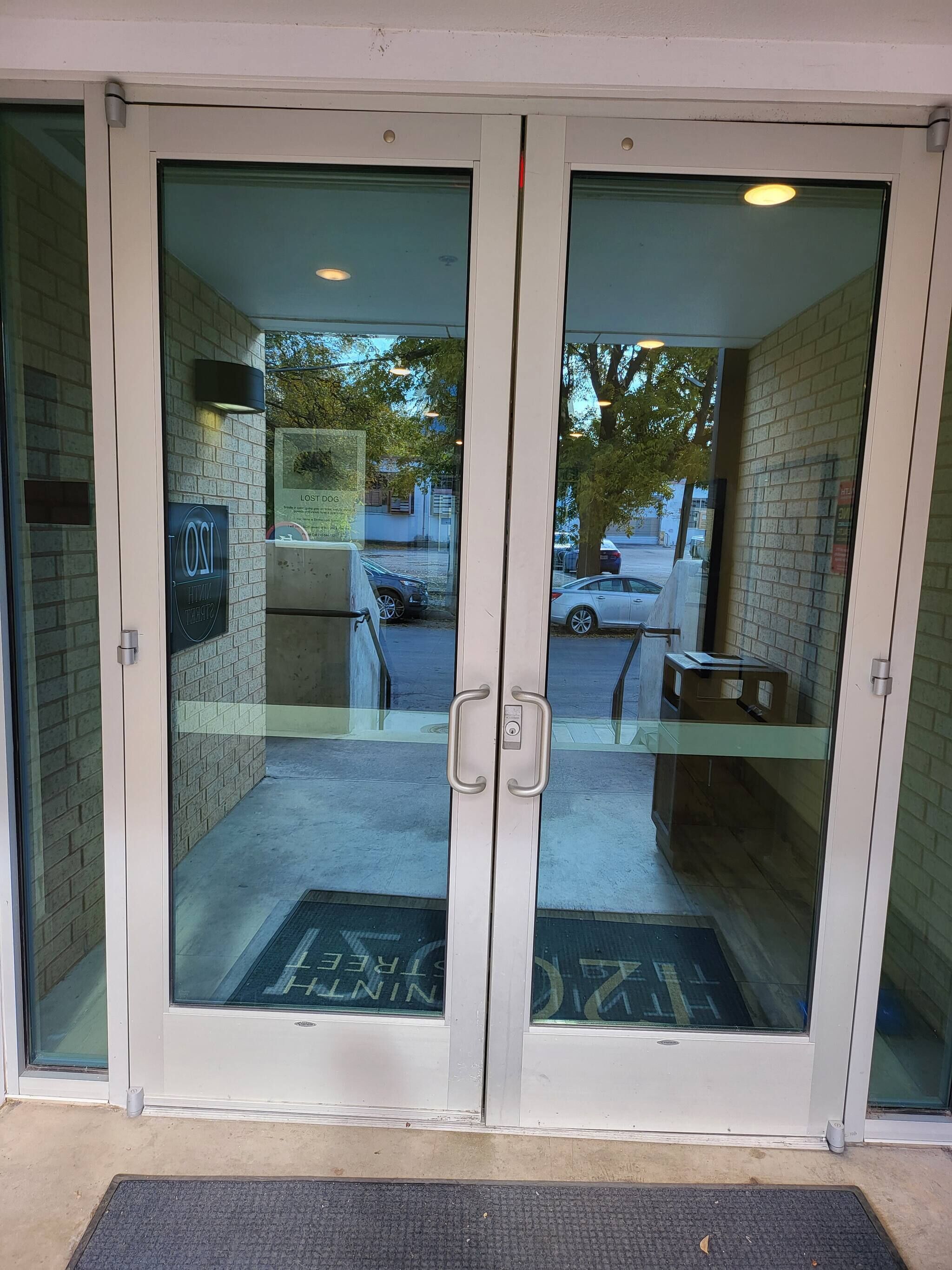 A double door entrance with glass panels and a metal handle, leading to a brick wall.