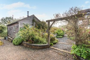 A rustic wooden house with a garden area, including a gravel path and various plants.