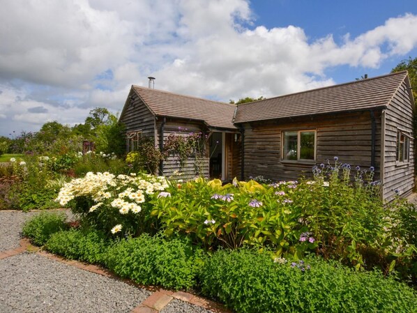 A wooden cabin with a sloped roof, surrounded by a garden with various flowers and shrubs.