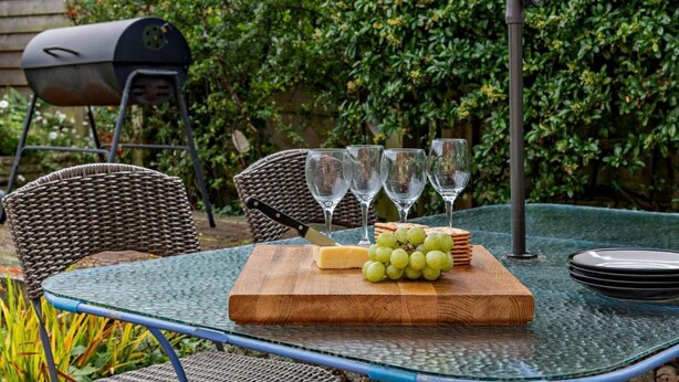 Een terras met rieten stoelen, een glazen tafel, een houten snijplank met kaas en druiven, en een barbecue op de achtergrond.