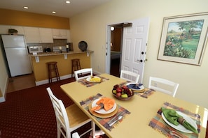 Un coin repas avec une table dressée pour quatre personnes, une cuisine en arrière-plan et une porte donnant sur une autre pièce.