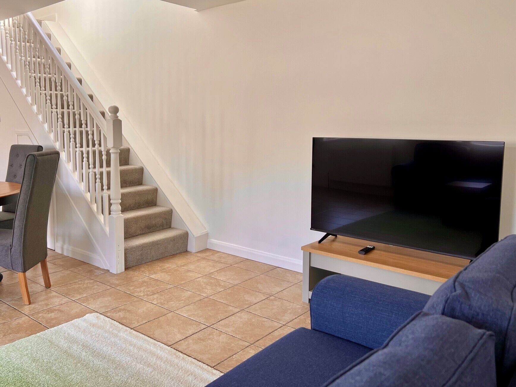A living room with a staircase, a flat-screen TV on a wooden stand, a blue sofa, and a dining area with chairs.
