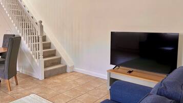 A living room with a staircase, a flat-screen TV on a wooden stand, a blue sofa, and a dining area with chairs.