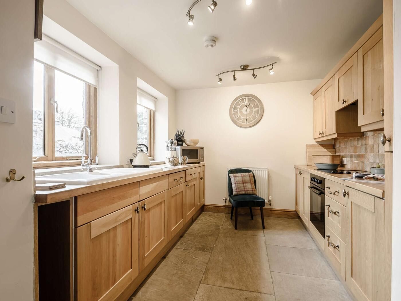 A kitchen with wooden cabinets, a sink, a kettle, a microwave, a clock, and a chair.