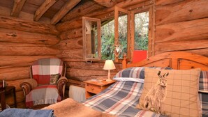 Ein gemütliches Blockhaus-Zimmer mit Bett, Sessel, Tisch und Fenster mit Blick auf Grünflächen.