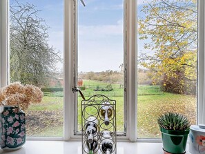 A window with a view of a rural landscape, a metal wine rack with bottles, a floral vase, and a potted plant. - 3 Bed in Stratford -upon- Avon (oc-54081) (Ettington)