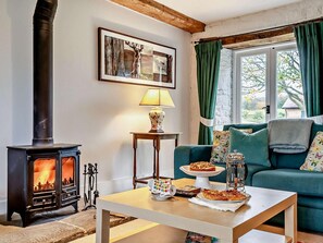 A cozy living room with a wood-burning stove, a sofa, a table with food, and a window with curtains. - 3 Bed in Stratford -upon- Avon (oc-54081) (Ettington)
