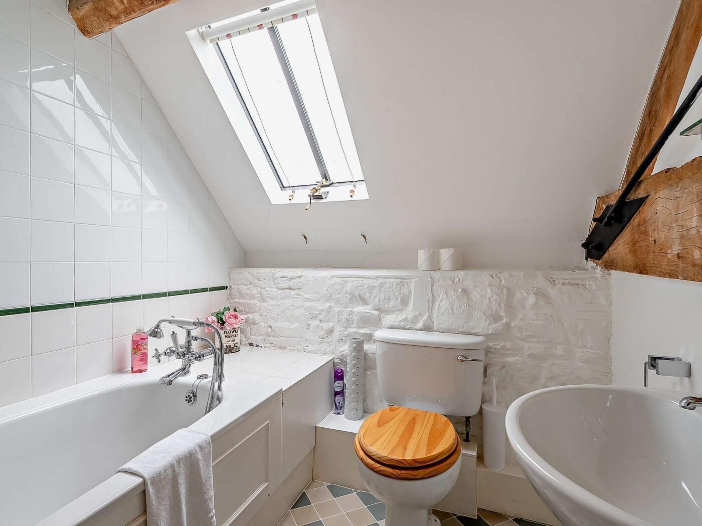 Un baño con bañera, inodoro y lavamanos, con azulejos blancos, tapa de inodoro de madera y tragaluz.
