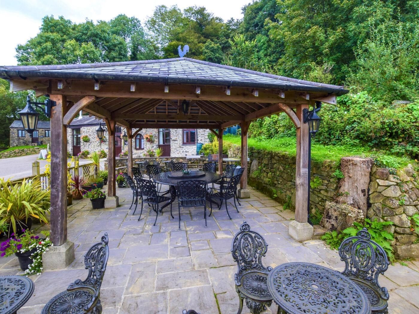 A covered outdoor seating area with stone walls, a wooden pergola, and a stone building in the background.