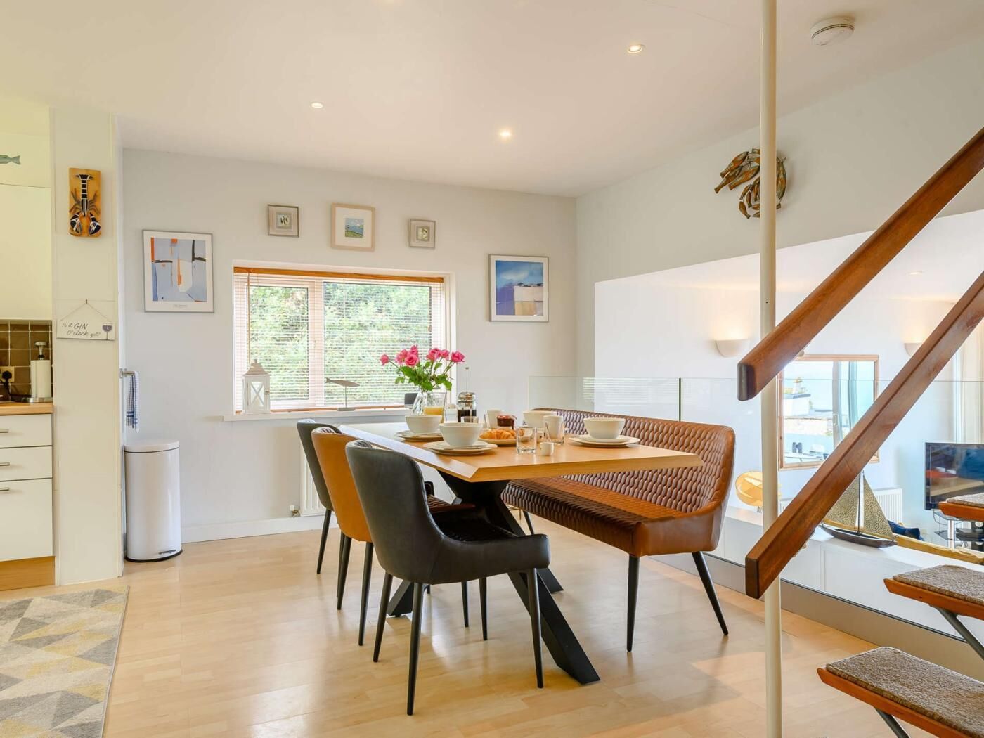 Un coin repas avec une table dressée pour quatre personnes, des chaises, une fenêtre avec des stores et un escalier.