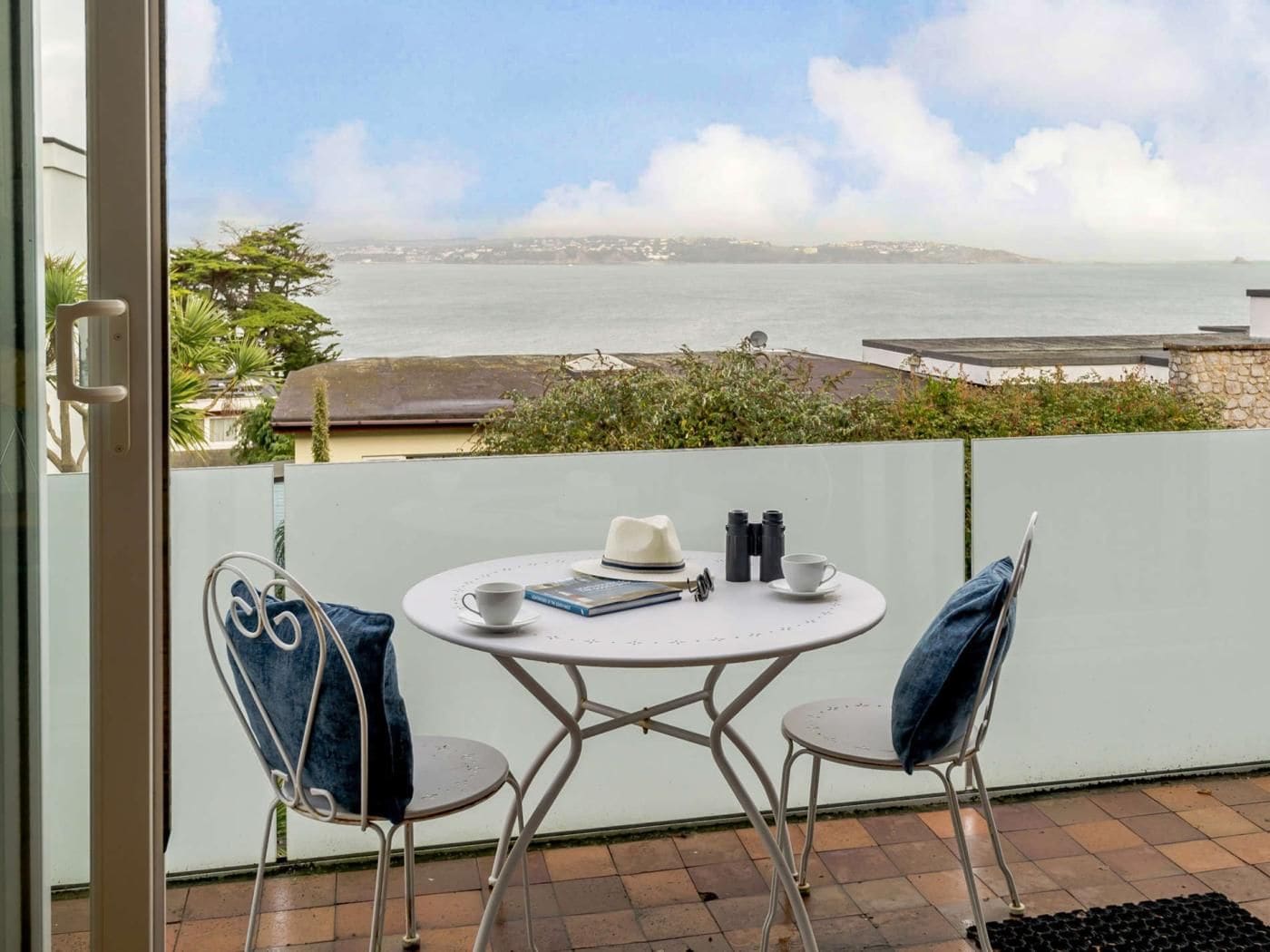 Un balcón con mesa y sillas, con vista a una masa de agua y una costa lejana.