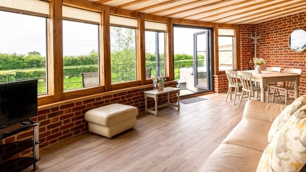 Amplia habitación con pared de ladrillo, ventanas grandes, techo de madera, un sofá, televisión y zona de comedor.