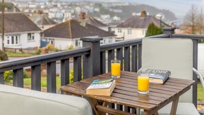 Ein Holztisch mit einem Buch und zwei Gläsern Orangensaft, ein weißer Stuhl und eine Ansicht von Häusern im Hintergrund.