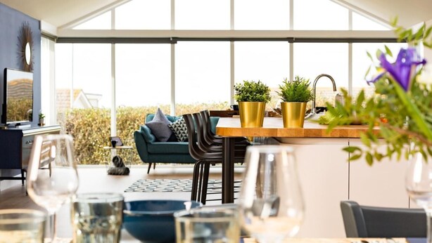 Une cuisine moderne dotée d’une grande fenêtre, un coin repas avec une table dressée pour deux personnes et un espace salon confortable avec un canapé et des fauteuils.