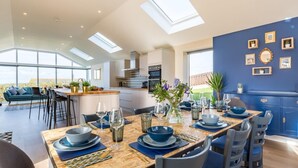 Un espace repas moderne avec une table en bois dressée pour quatre personnes, une cuisine en arrière-plan et de grandes fenêtres.