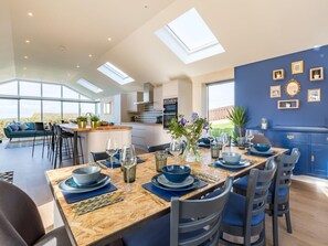A modern dining area with a wooden table set for four, kitchen in the background, and large windows. - 4 Bed in Bideford (oc-54448) (Appledore)