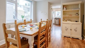A dining area with a wooden table and chairs, a kitchen in the background, and a white cabinet with floral designs. - 3 Bed in Brixham (oc-bx001) (Brixham)