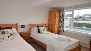 A bedroom with two beds, a wooden wardrobe, a window with a view of buildings, and a radiator. - 3 Bed in Brixham (oc-bx018) (Brixham)