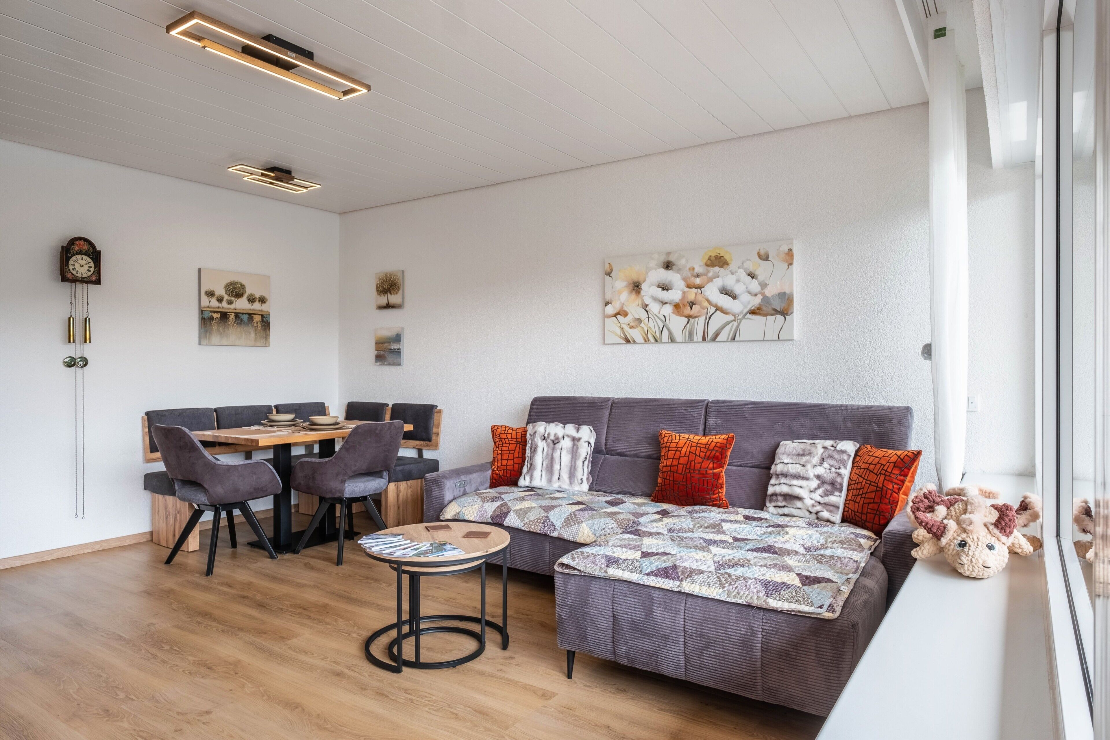 Un salon moderne avec un canapé, une table à manger et des chaises, ainsi qu’une table basse. On y trouve également des tableaux accrochés au mur et une horloge.
