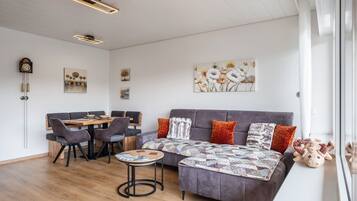 Un salon moderne avec un canapé, une table à manger et des chaises, ainsi qu’une table basse. On y trouve également des tableaux accrochés au mur et une horloge.