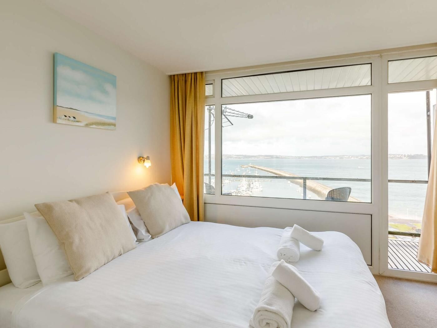 A hotel room with a bed, a painting, a window with a view of the sea, and a curtain.