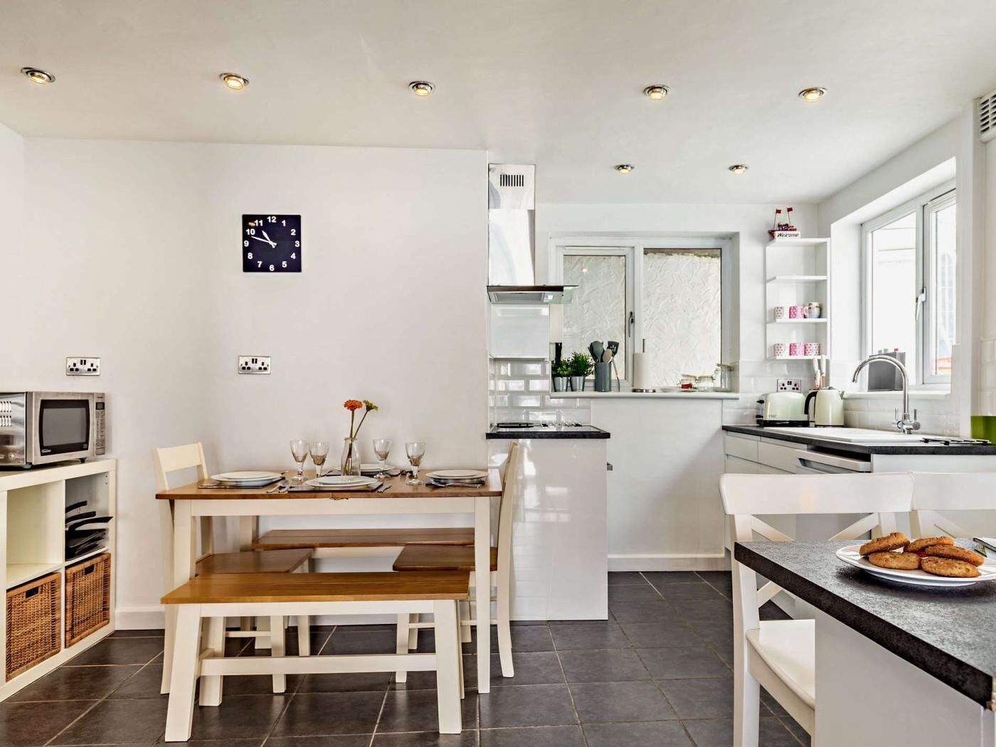 A modern kitchen with white cabinetry, a dining table with chairs, a clock on the wall, and a window with a view.