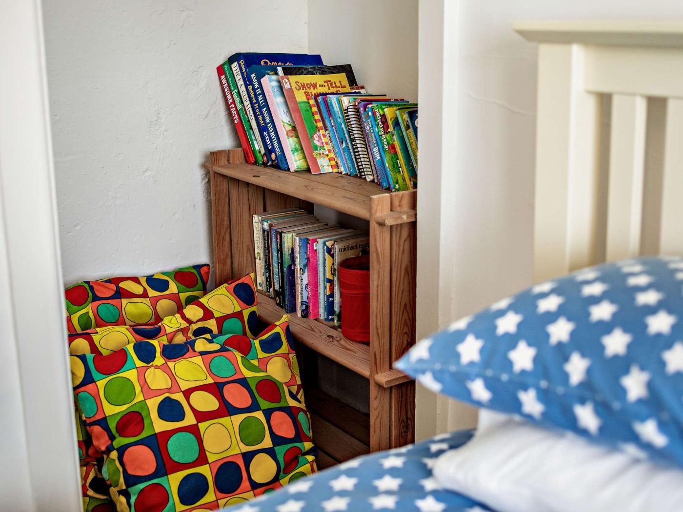 Ein Bücherregal mit Büchern und einem farbenfrohen Kissen in einem Zimmer mit weißer Wand und einem Kinderbett mit einem blau-weißen Kissen.