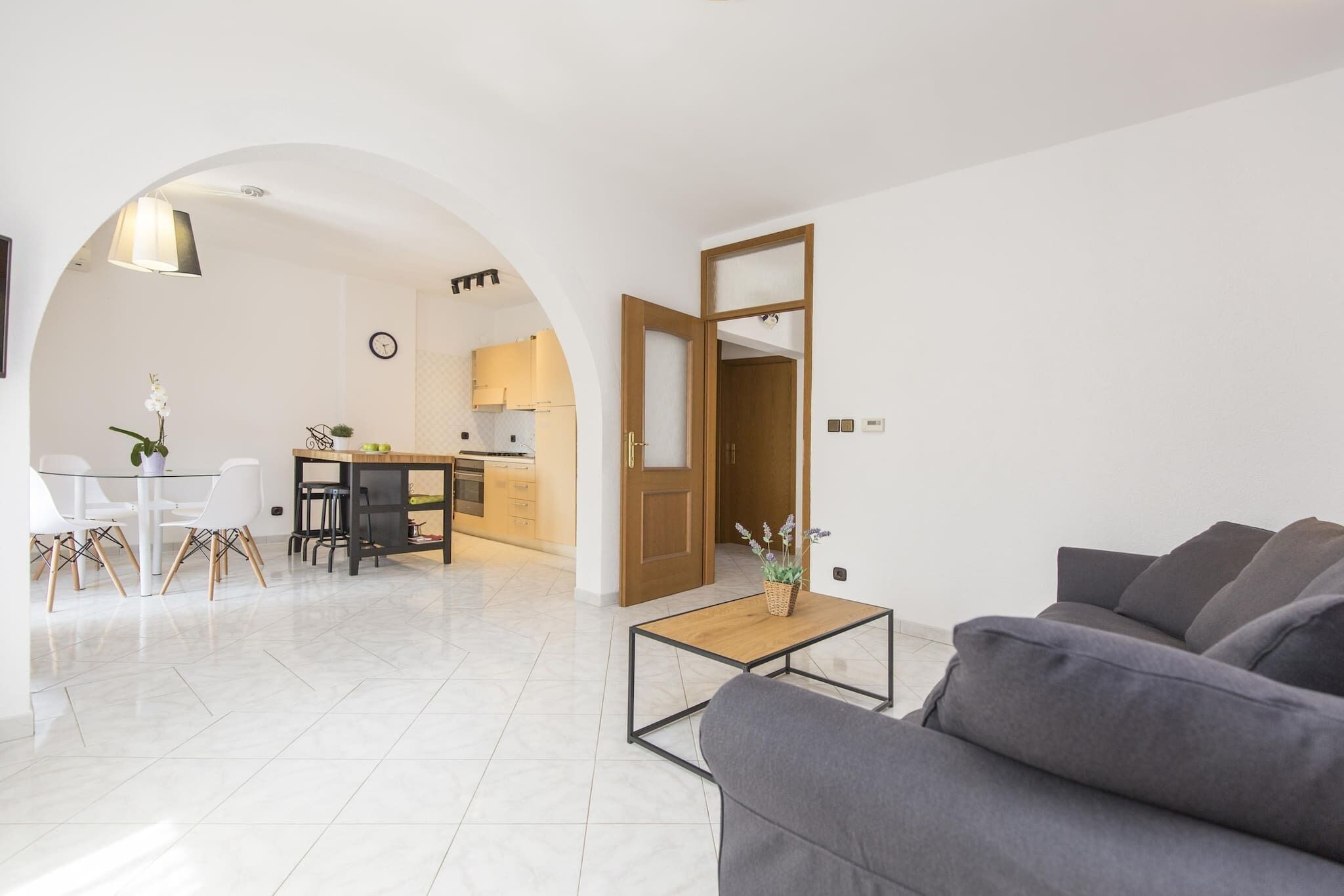 Ein modernes Wohnzimmer mit einem grauen Sofa, einem Holztisch und einem Essbereich mit weißen Stühlen sowie einer kleinen Küche im Hintergrund.