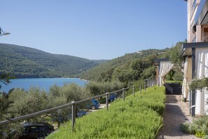 Blick von einer Hügelkuppe auf ein Gebäude, im Hintergrund ein klarblauer See, ein Kiesweg und geparkte Autos.