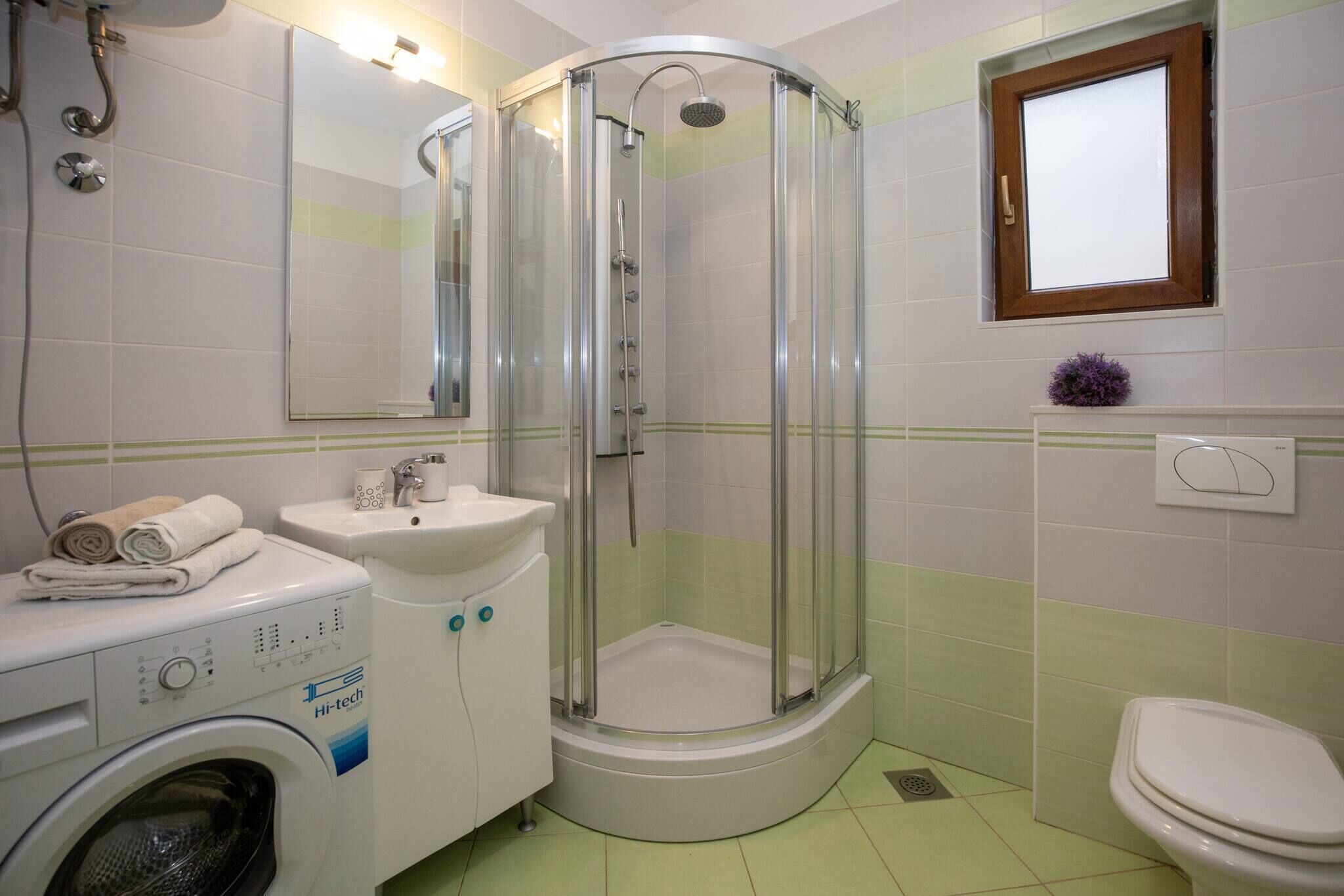Casa de banho com máquina de lavar roupa, pia, sanita e cabine de duche em vidro.