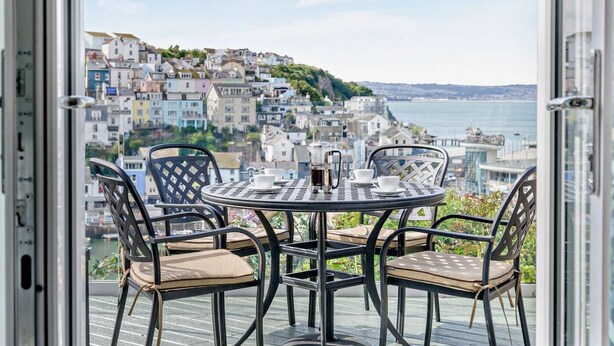 Ein Balkon mit rundem Tisch und Stühlen, Blick auf eine Küstenstadt mit bunten Gebäuden.