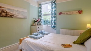 Un dormitorio con cama, una mesita de noche con lámpara, una ventana con cortinas, un radiador y una pared con un cuadro y un cartel.