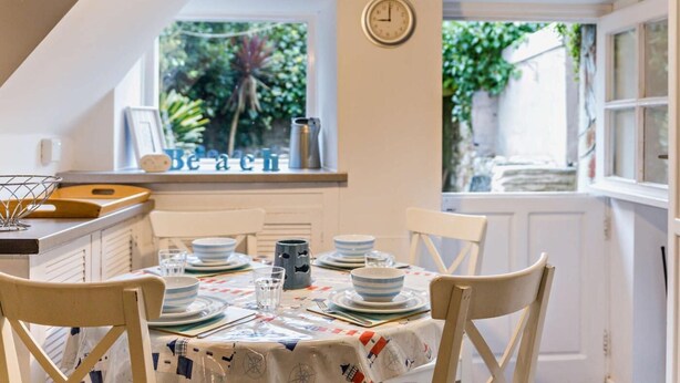 Un comedor con una mesa dispuesta para cuatro, un reloj en la pared y una ventana con vistas a zonas verdes.