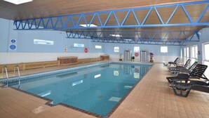 Ein Hallenbad mit Holzbalkendecke, blauen Sicherheitsgeländern und Liegestühlen am Rand.