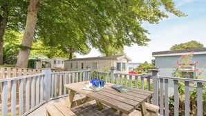 Eine Holzterrasse mit Tisch und Bänken, umgeben von einem weißen Zaun, mit Blick auf Mobilheime und ein Gewässer im Hintergrund.