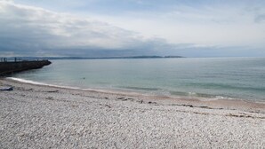 En strand med småsten, ett lugnt hav och en kustlinje som ligger en bit bort.