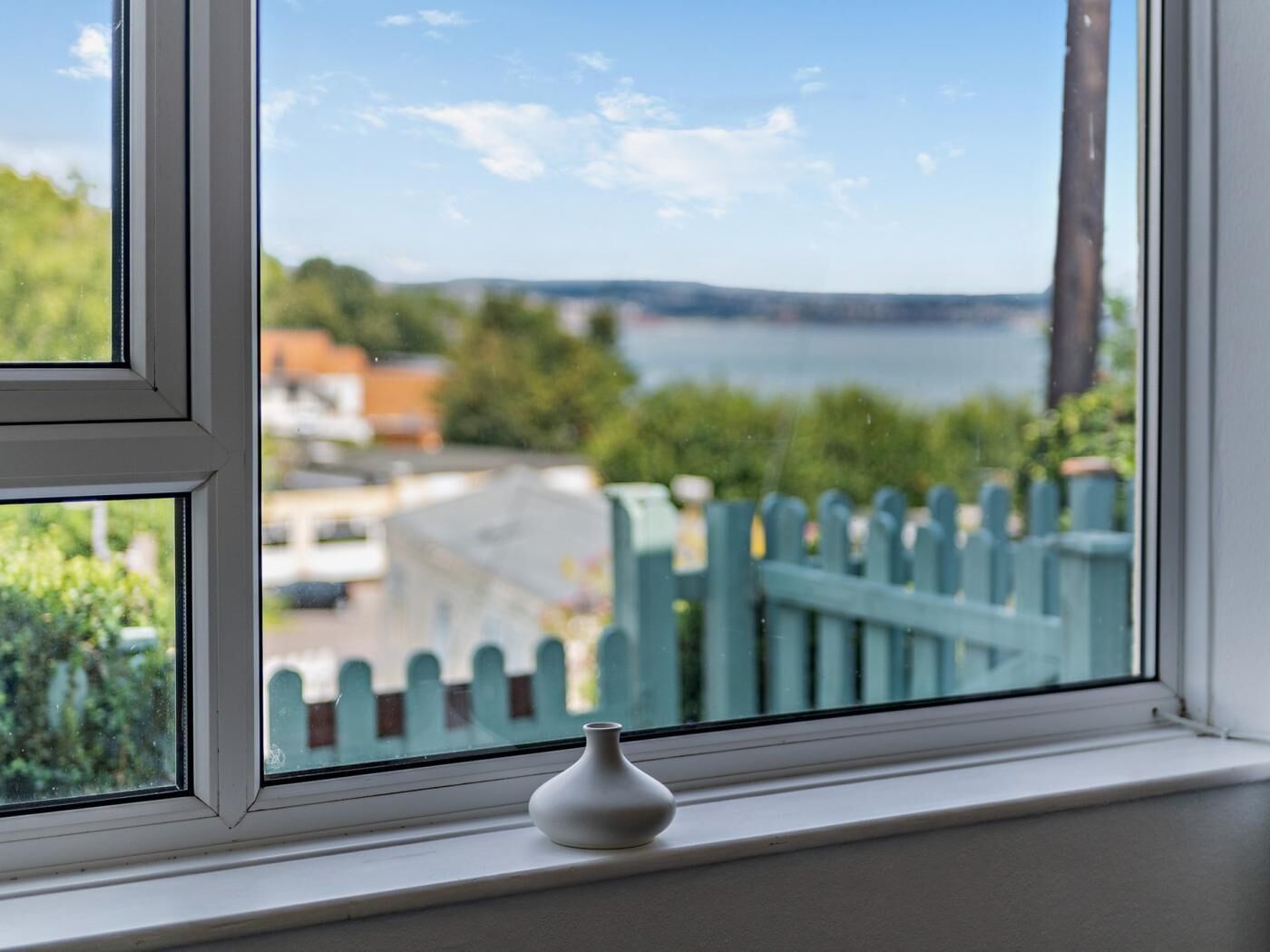 A window with a white vase on the sill, a view of a body of water, and a green fence in the background.
